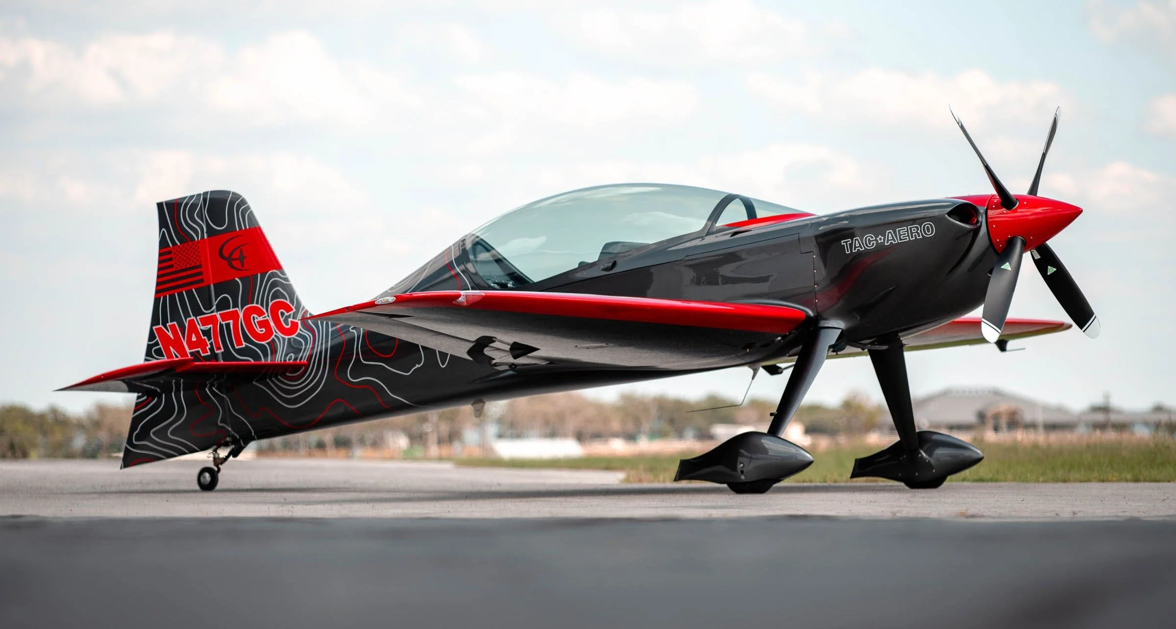 A small, black and red single-engine aircraft with a sleek design and a propeller in front, parked on the tarmac under a cloudy sky.