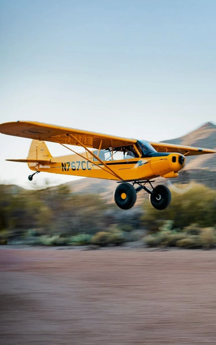 A yellow small aircraft flying low over desert terrain with mountains in the background.