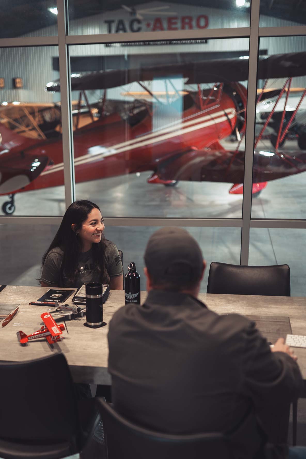 Two people seated at a table inside an aviation hangar, with a red biplane aircraft and the sign 'TAC AERO' visible through the window behind them.