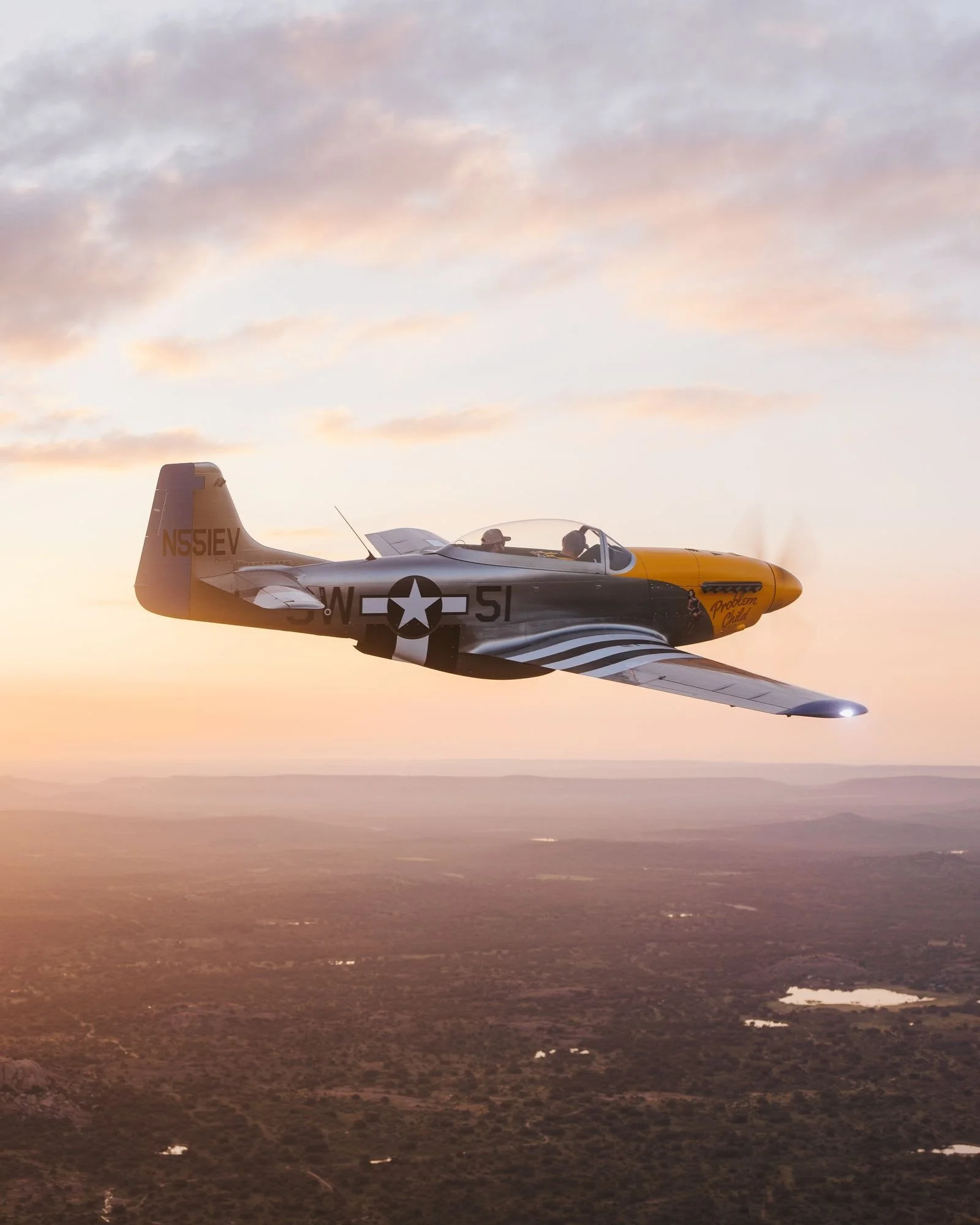 A vintage military aircraft flying at sunset over a landscape with lakes and trees.