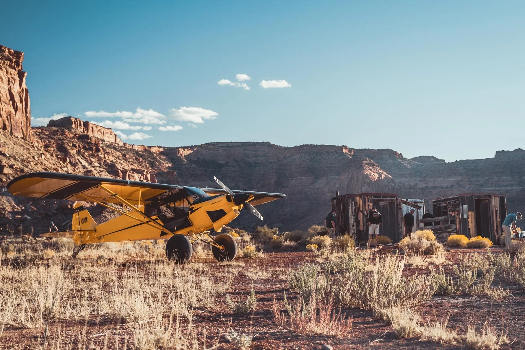 A yellow small airplane parked on a desert terrain with dry bushes, with a wrecked train car and rocky hills in the background under a blue sky with a few clouds.