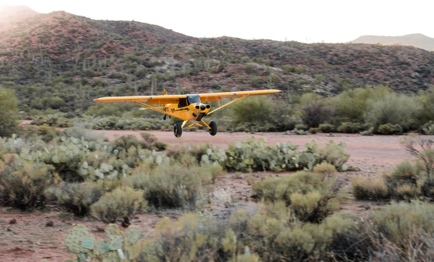 A yellow aircraft flying low over desert landscape with cacti and mountains in the background.
