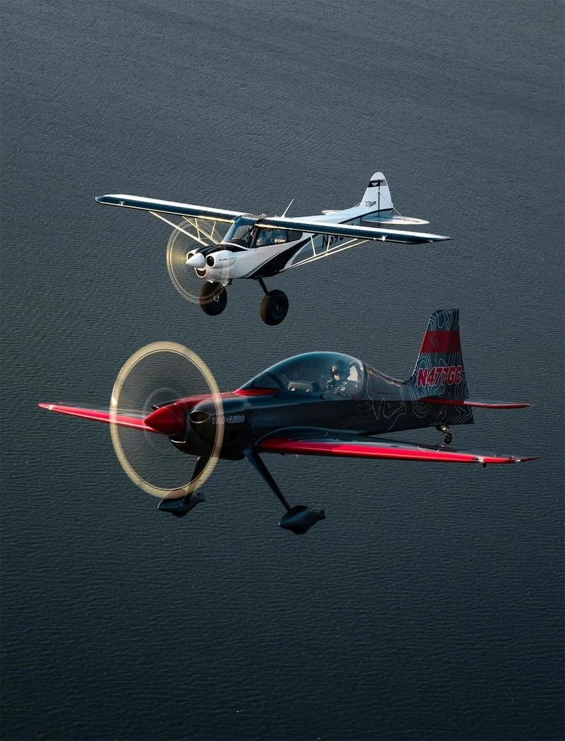 Two small planes flying over water, with the bottom plane in black and red and the top plane in white and black.