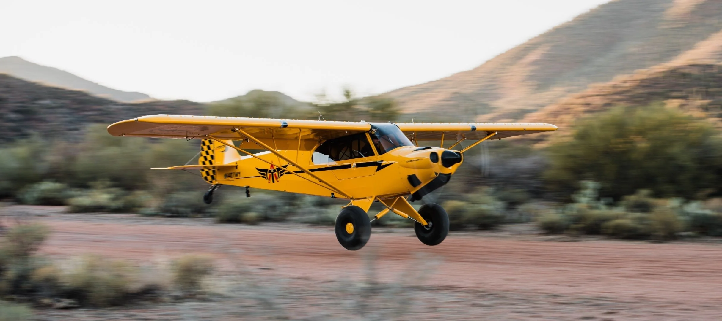 Yellow single-engine airplane flying over desert landscape with hills and sparse vegetation.
