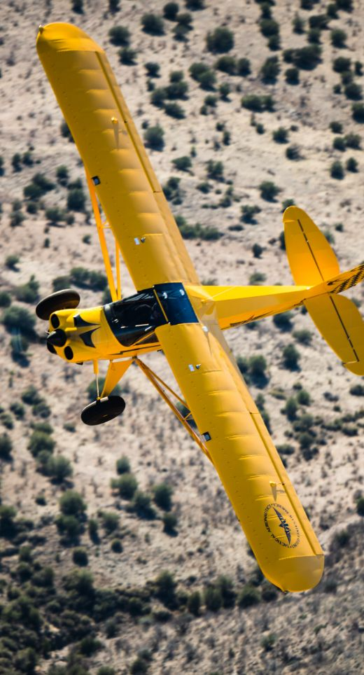 A yellow aircraft flying low over a desert landscape with sparse bushes.
