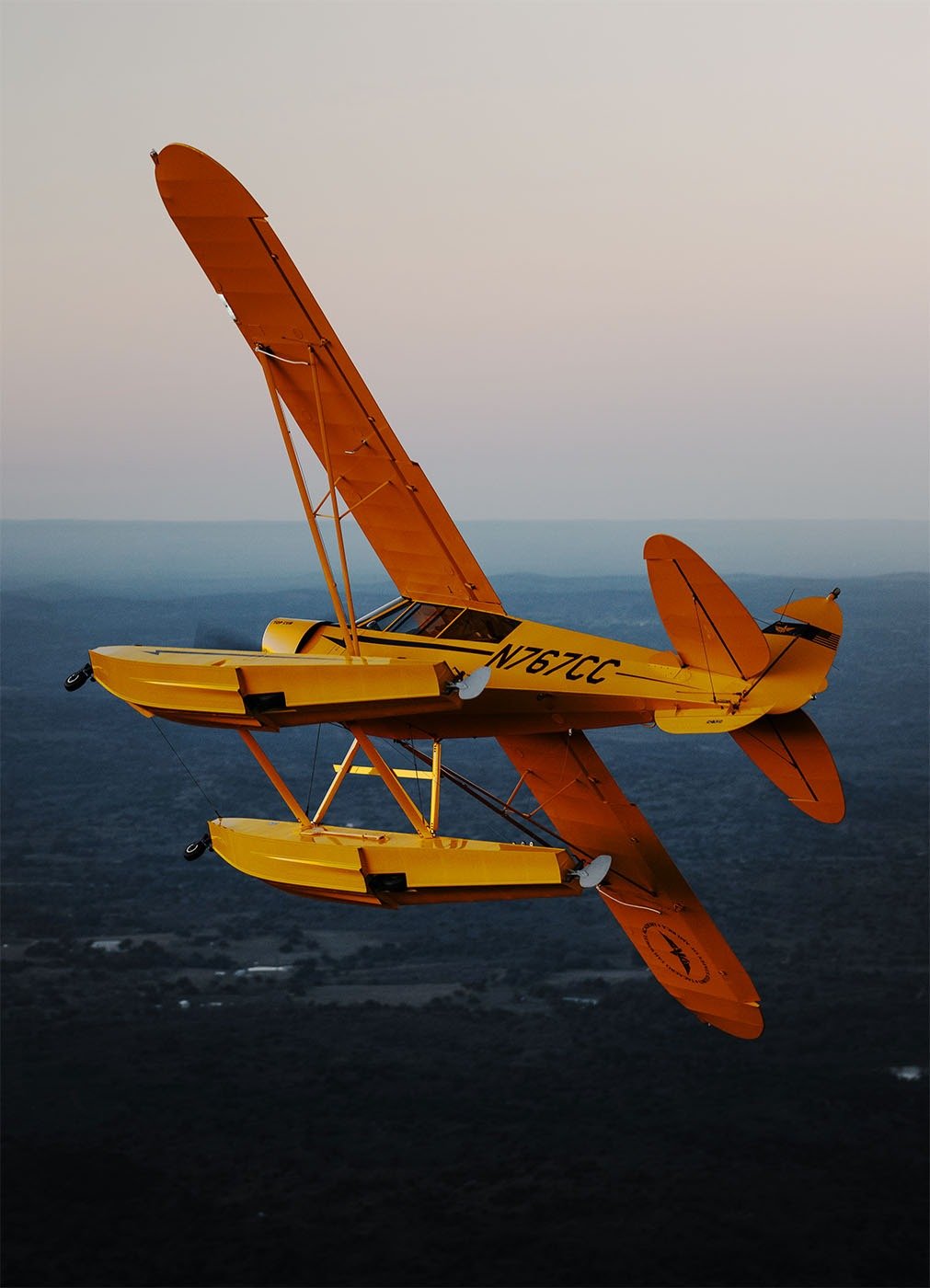 A yellow and orange seaplane flying above a landscape with mountains and fields, reflected in the water surface.