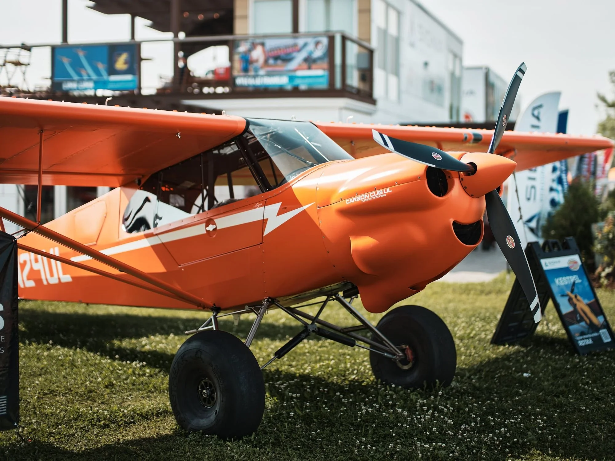 Orange single-engine propeller airplane on display outdoors, with a building and trees in the background.
