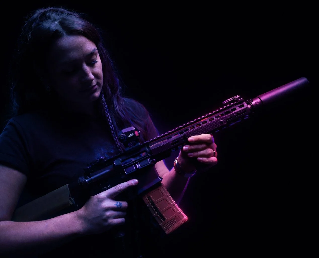 A woman holds a rifle with a silencer and a mounted scope, illuminated by purple and pink lighting in a dark background.