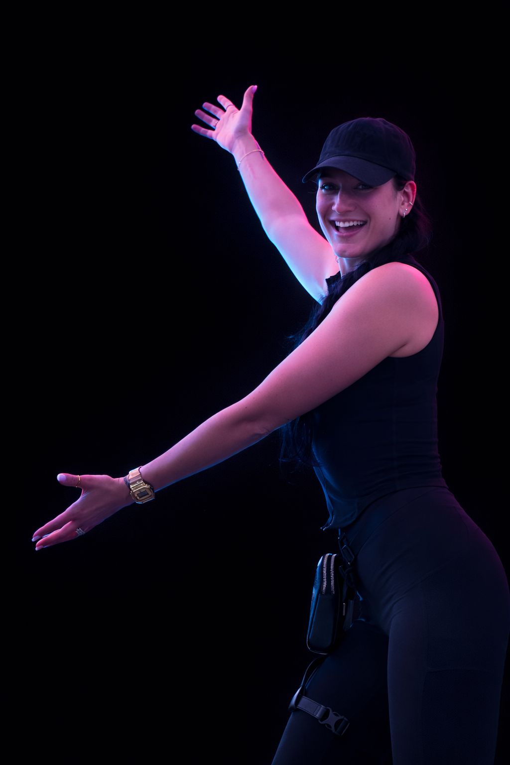 A woman wearing a black cap and sleeveless black top, smiling, with one arm raised and the other extended, against a black background with colorful lighting.