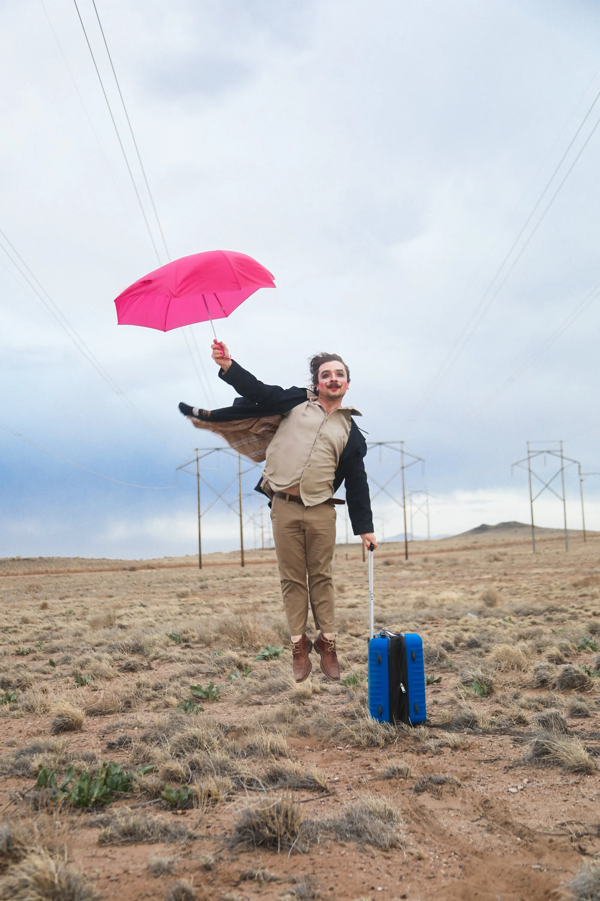 A person dressed as a clown, with makeup and a beard, jumping in the air with a suitcase at their feet, holding a pink umbrella in a barren landscape with power lines and cloudy sky in the background.