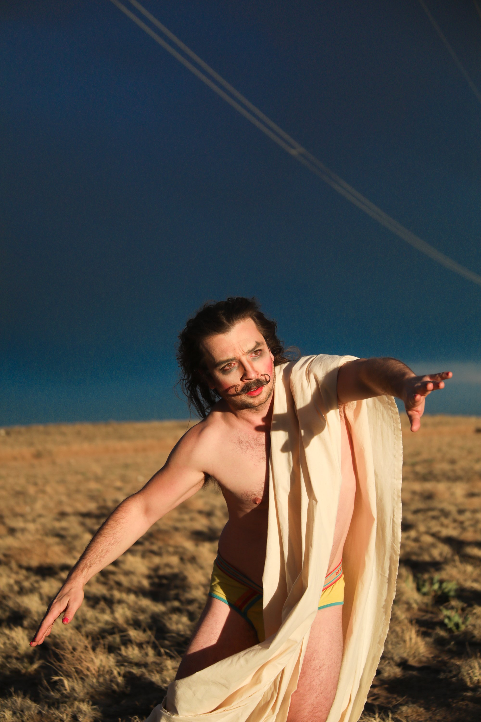 A man with a mustache and long hair, wearing only colorful underwear and a beige cloth, is stretching out his arm in a desert landscape under a dark sky with contrails.