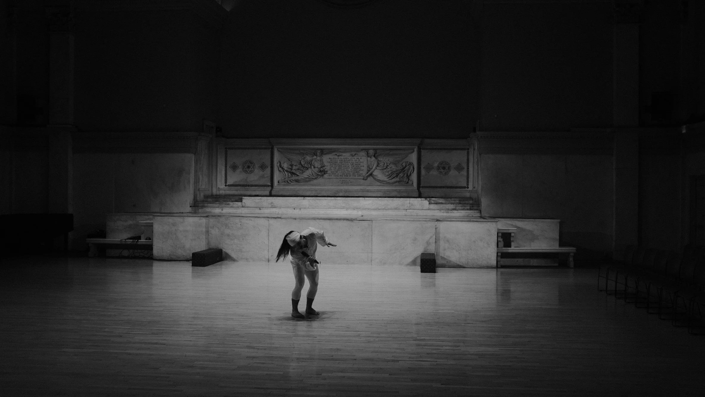A woman dancing alone in a large empty hall with wooden floors and decorative artwork on the back wall, illuminated by a spotlight.
