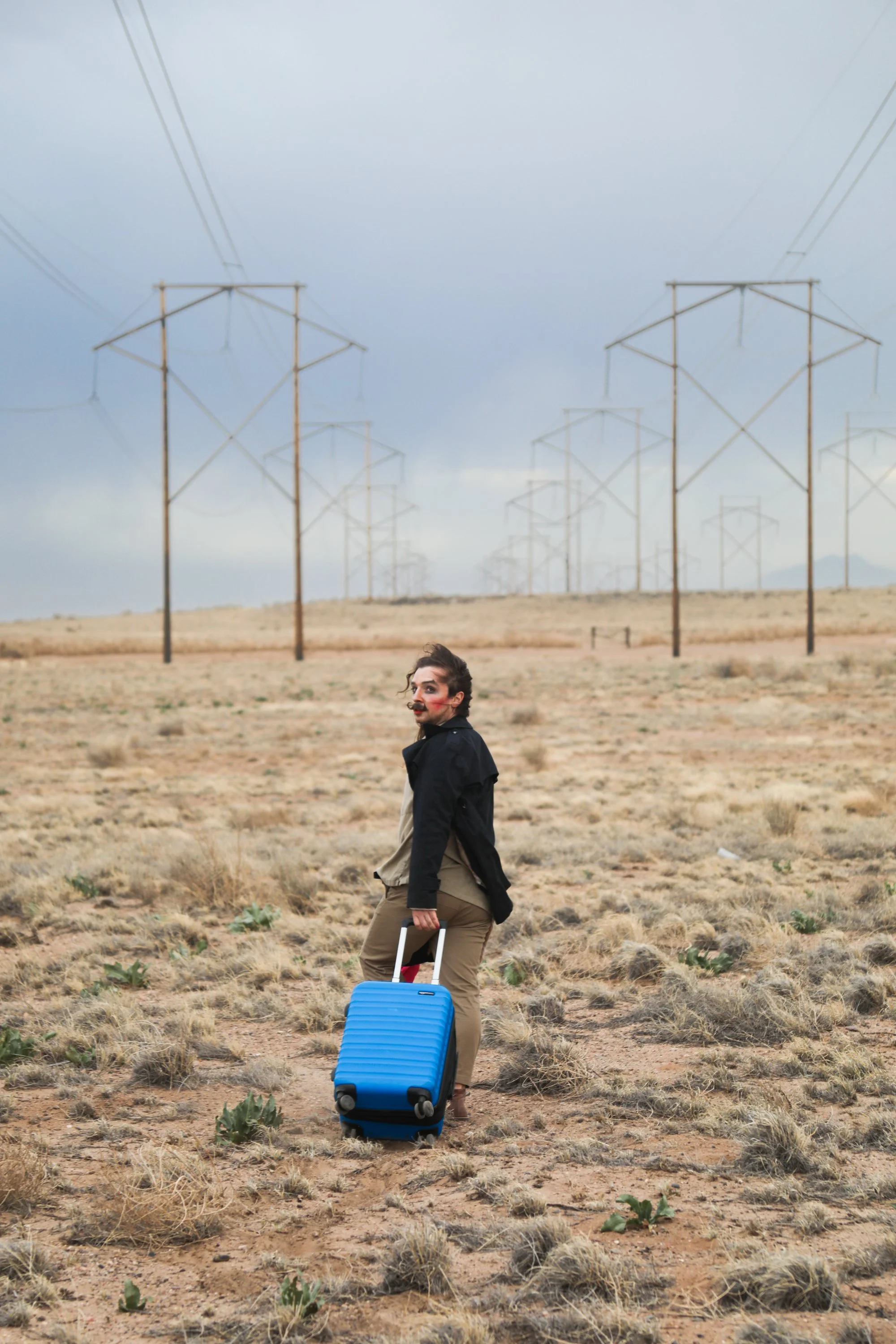 Person with brown hair, holding a blue rolling suitcase, standing in a barren desert landscape with power lines in the background under cloudy sky.