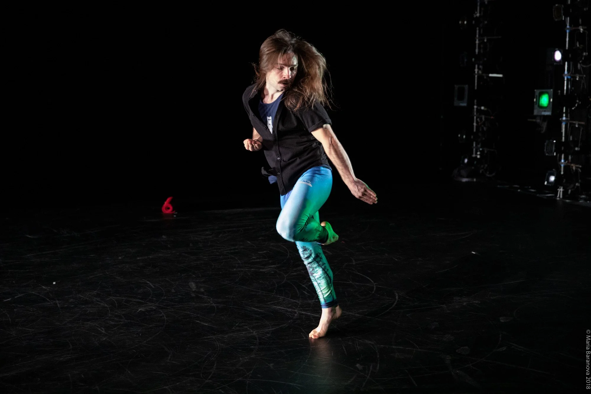 Person dancing on stage with black background, wearing colorful leggings and a black shirt.