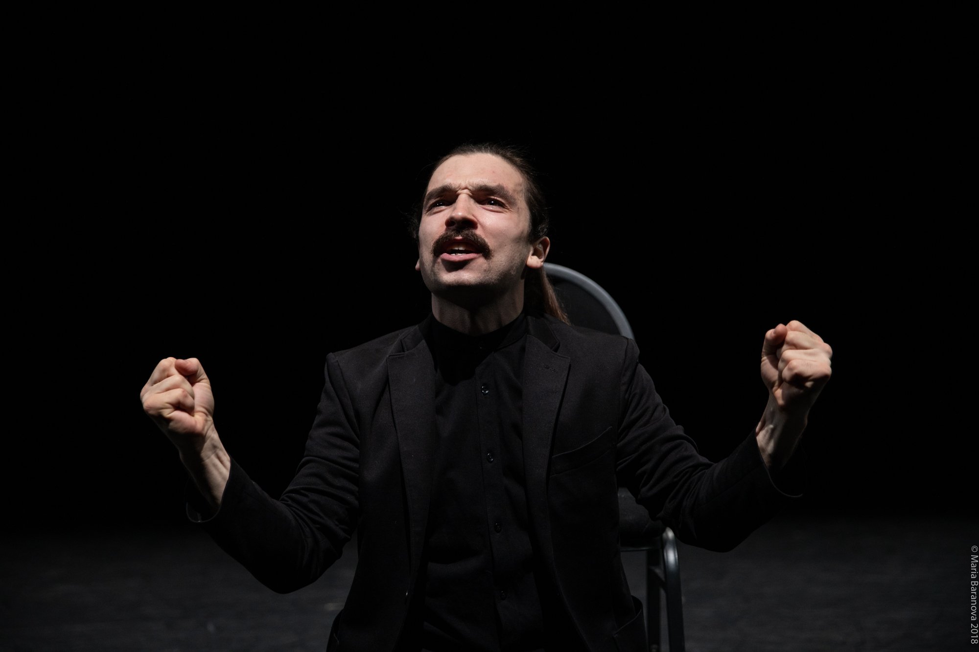 A man with dark hair and a mustache, dressed in black, is sitting on a chair with a dark background. He appears passionate, with his fists clenched and raised, and an intense facial expression.