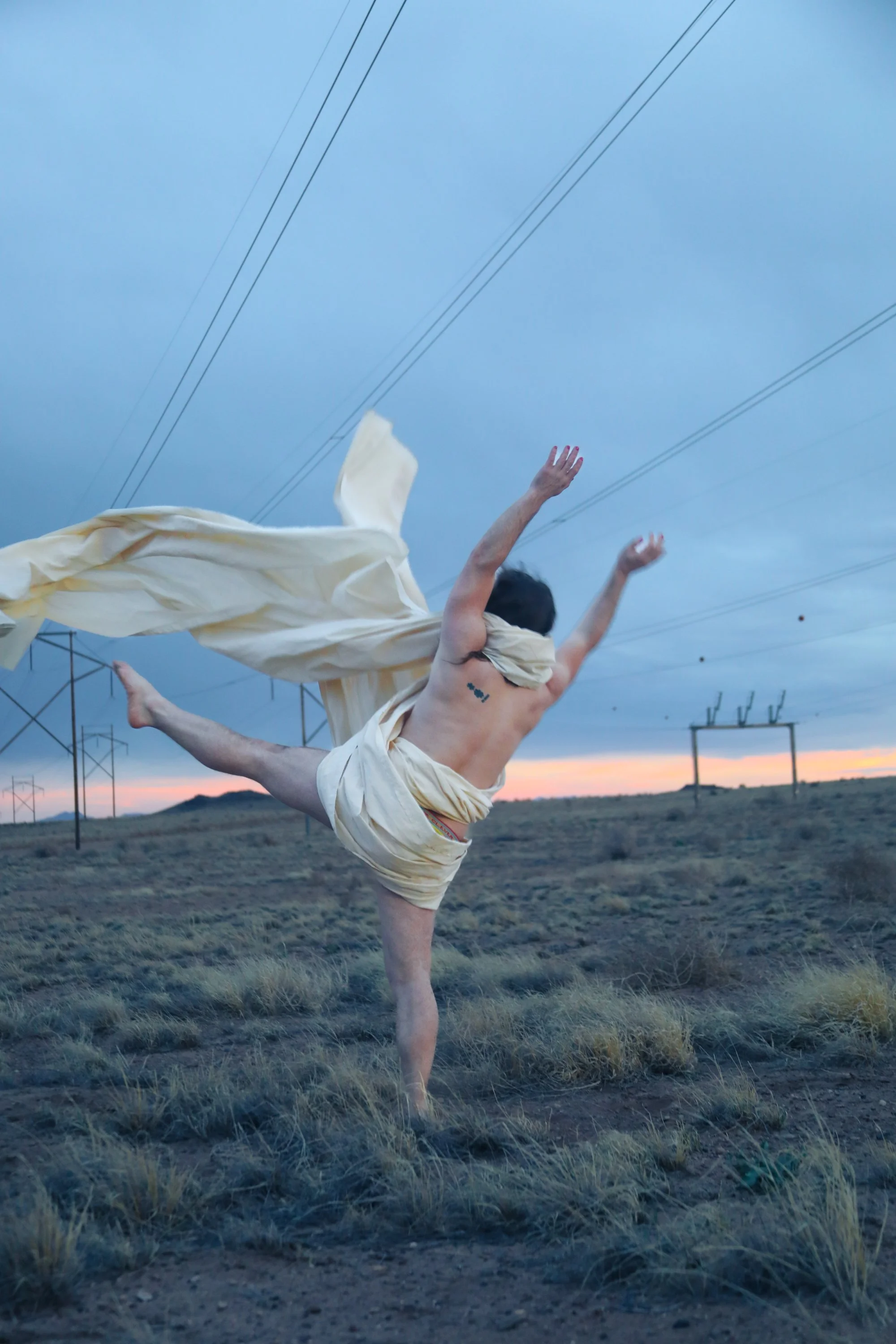 A shirtless man with a tattoo, wearing loose light-colored clothing, balancing on one hand in a desert landscape with power lines and a sunset in the background.