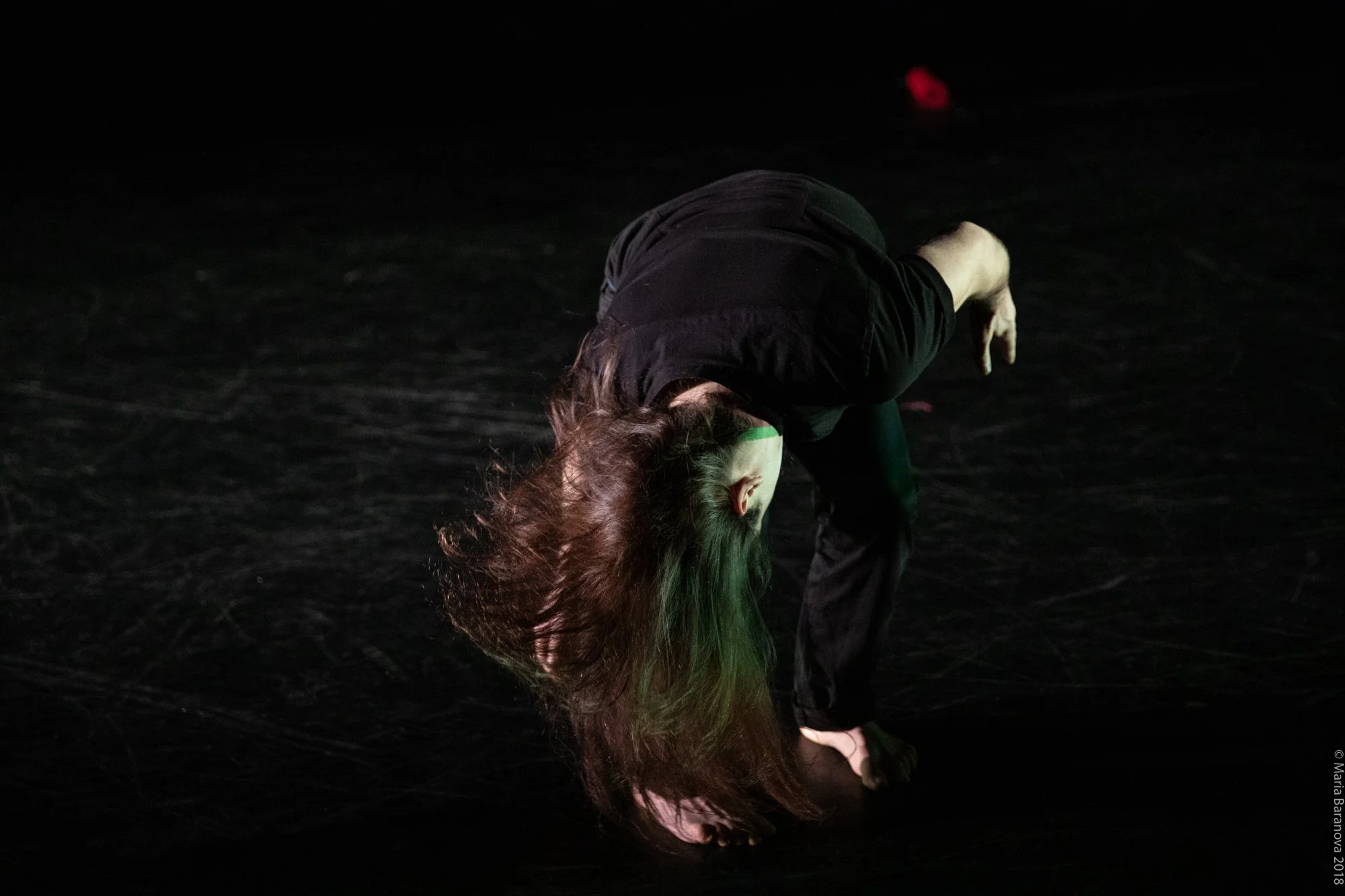 A person with long hair bent over in a dark setting, possibly performing or practicing, wearing a black shirt and pants.