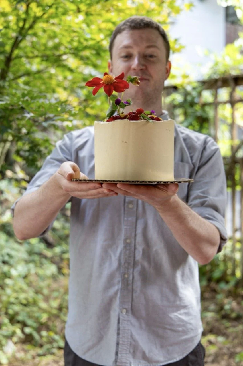 A man in a gray shirt holding a white cake decorated with colorful flowers outdoors.