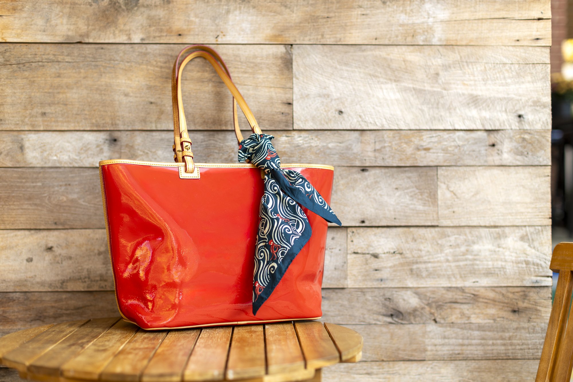A red handbag with tan handles and a blue patterned scarf tied to one handle, placed on a wooden table against a wooden wall background.