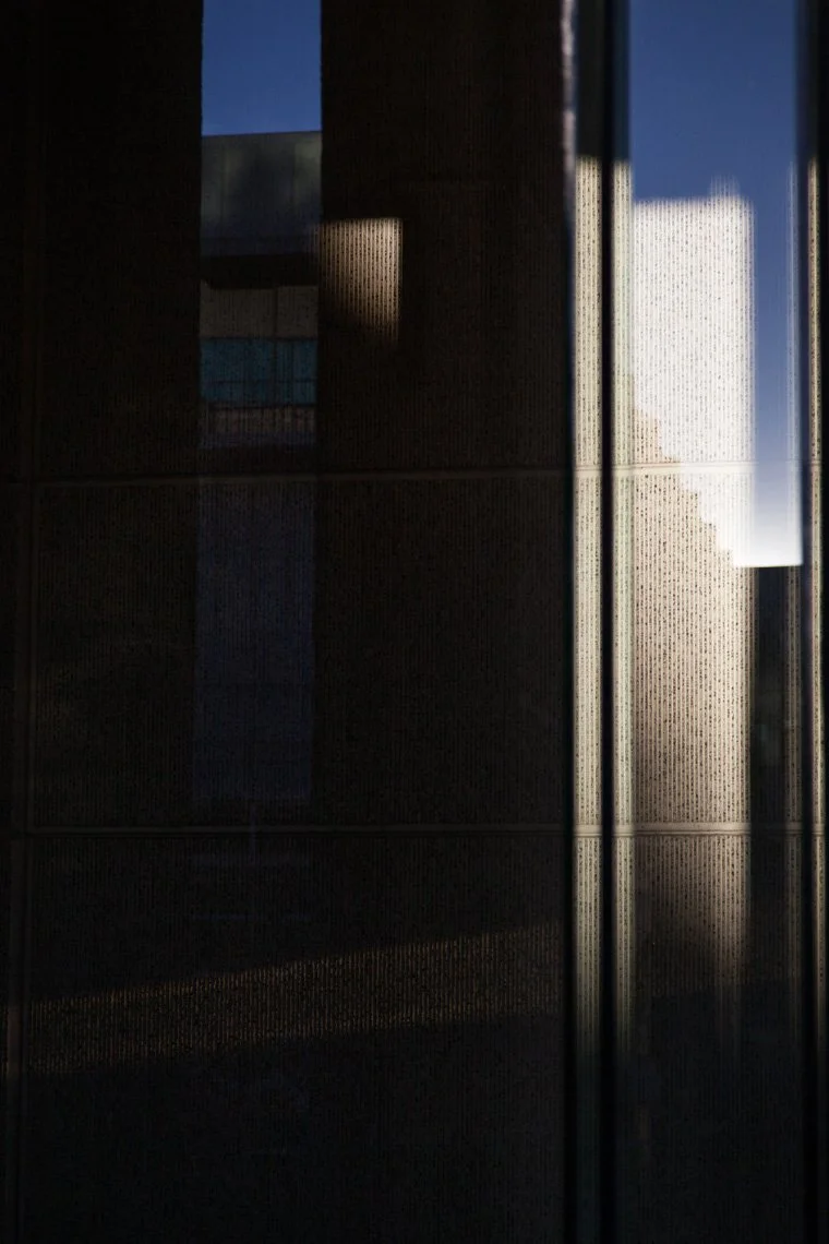 The image shows a dark interior with a large window or glass door. Sunlight streams through the window, casting bright reflections and shadows on the dark wall or panels inside.