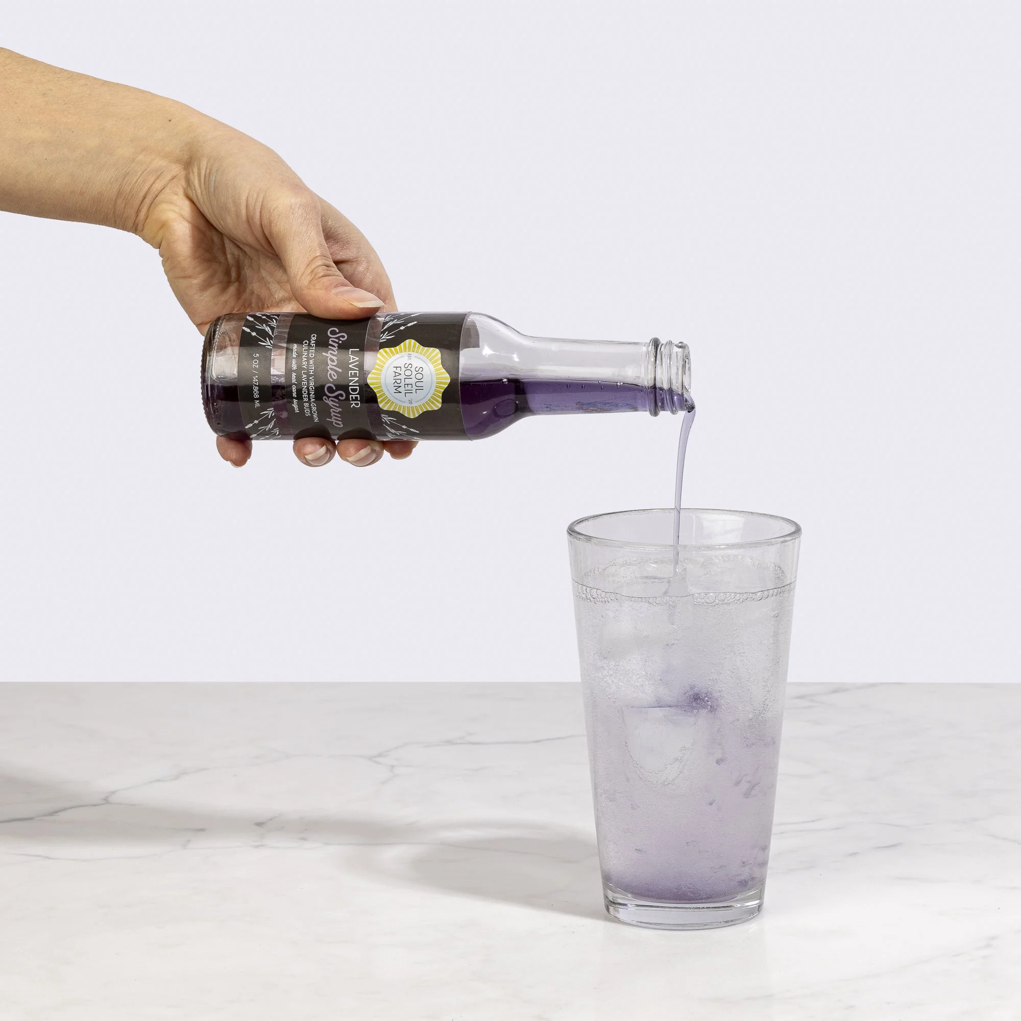 A hand holding a bottle of lavender syrup pouring into a glass of water, with a minimal background.