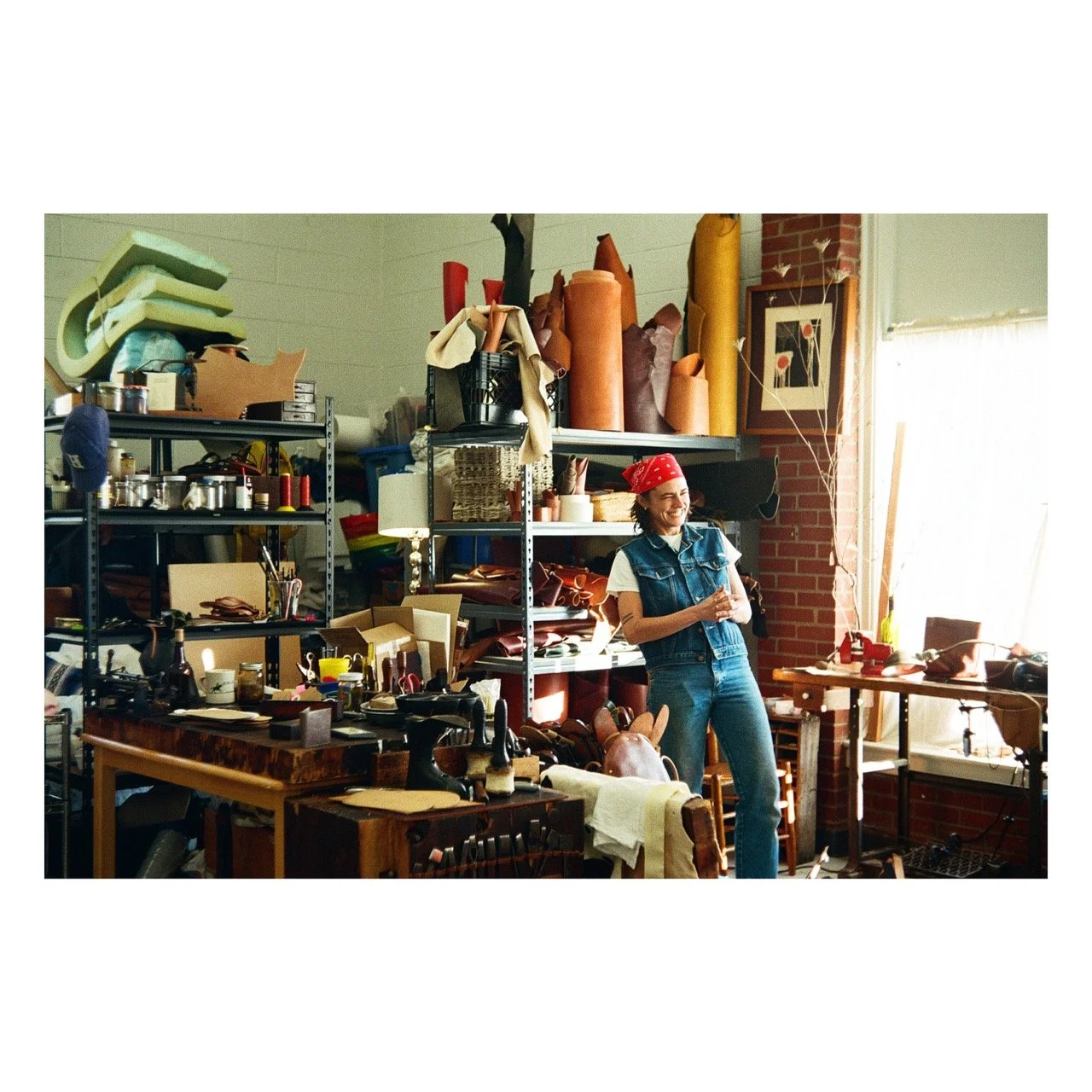 A smiling woman with a red bandana, white shirt, and denim vest stands in a cluttered workspace filled with crafting supplies, leather and fabric rolls, and tools. Shelves behind her hold various materials, and sunlight streams through a window on th