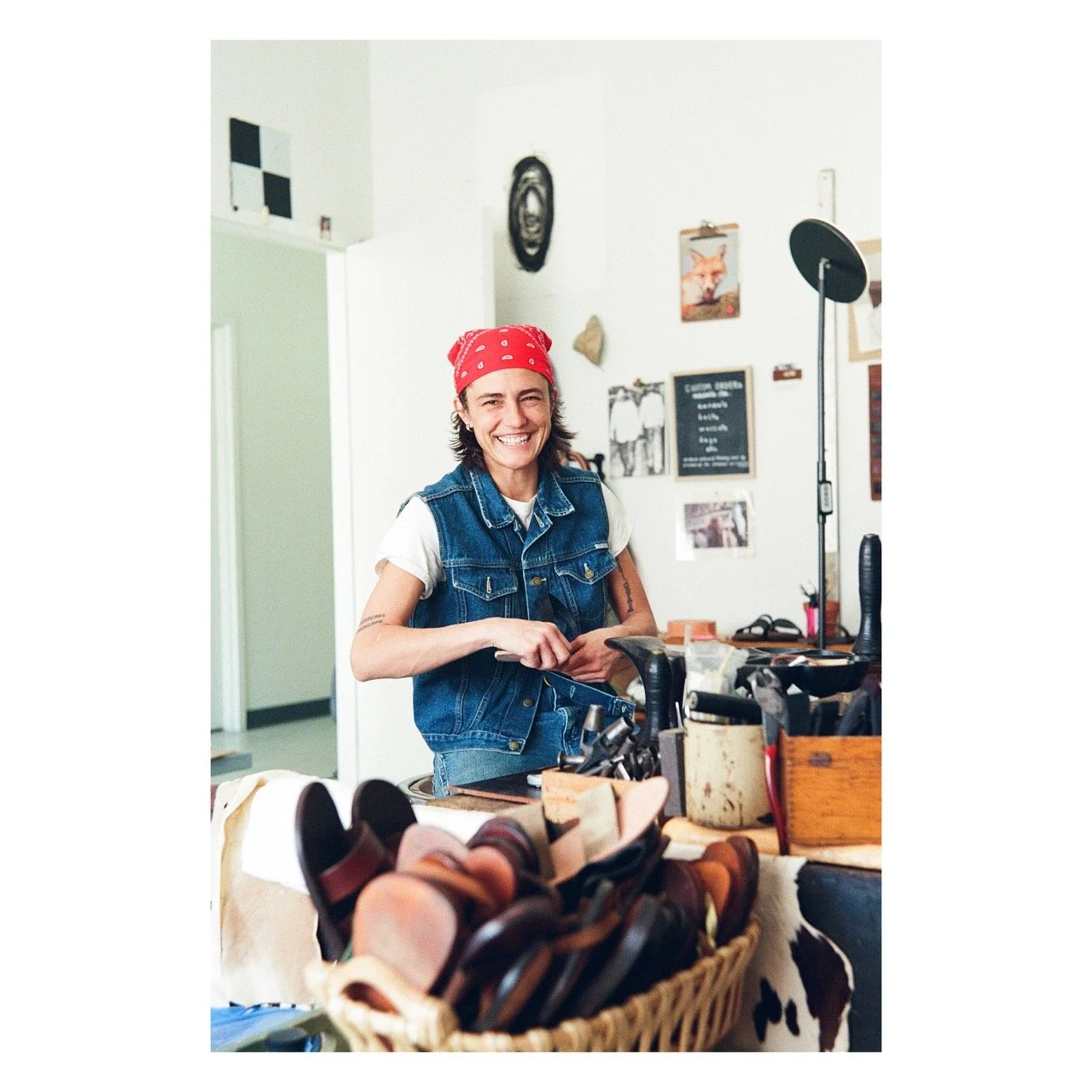A smiling woman with a red bandana, wearing a denim vest over a white t-shirt, standing behind a counter with sunglasses in a basket, inside a shop with eclectic decor including framed photos and a chalkboard menu.