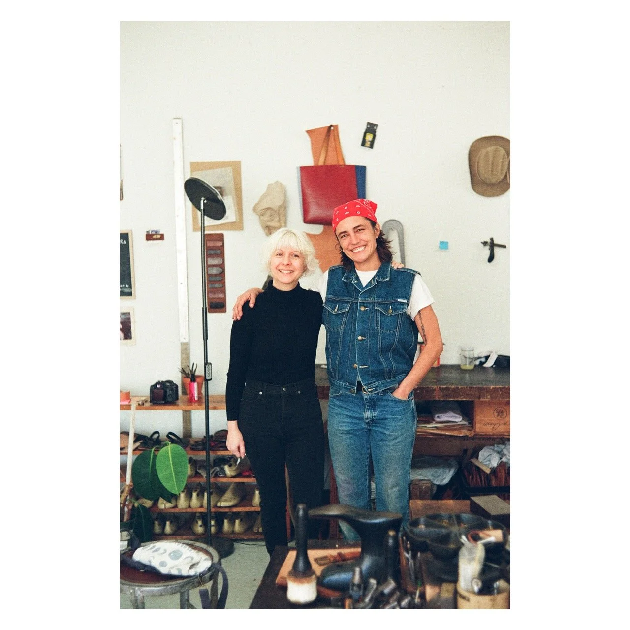 Two women standing in a workshop, smiling. One woman with short white hair wearing a black turtleneck, the other with long dark hair and a red bandana, wearing a denim vest and jeans. The workshop background includes shoes, tools, and hanging bags.