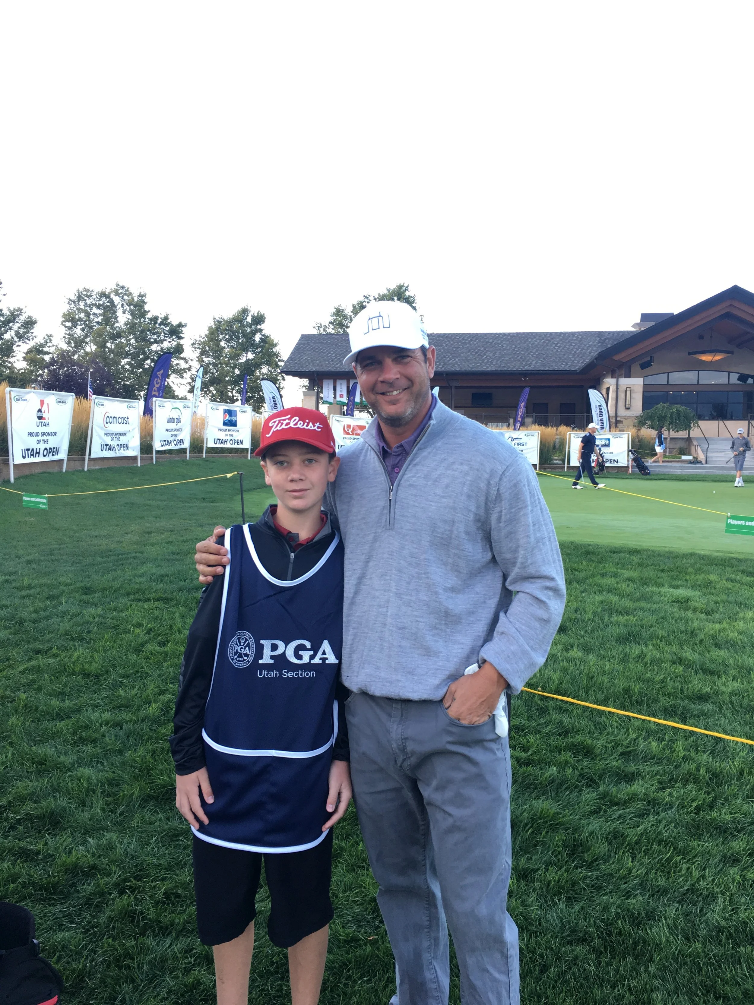 A man and a boy standing together outdoors at a golf event, with golf signage and people playing in the background.