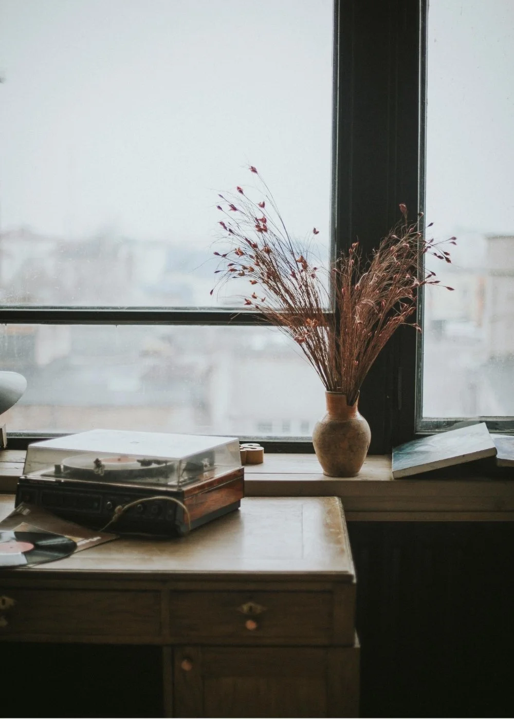 A vintage record player on a wooden desk beside a vase with dried flowers, set against a large window showing an overcast cityscape.