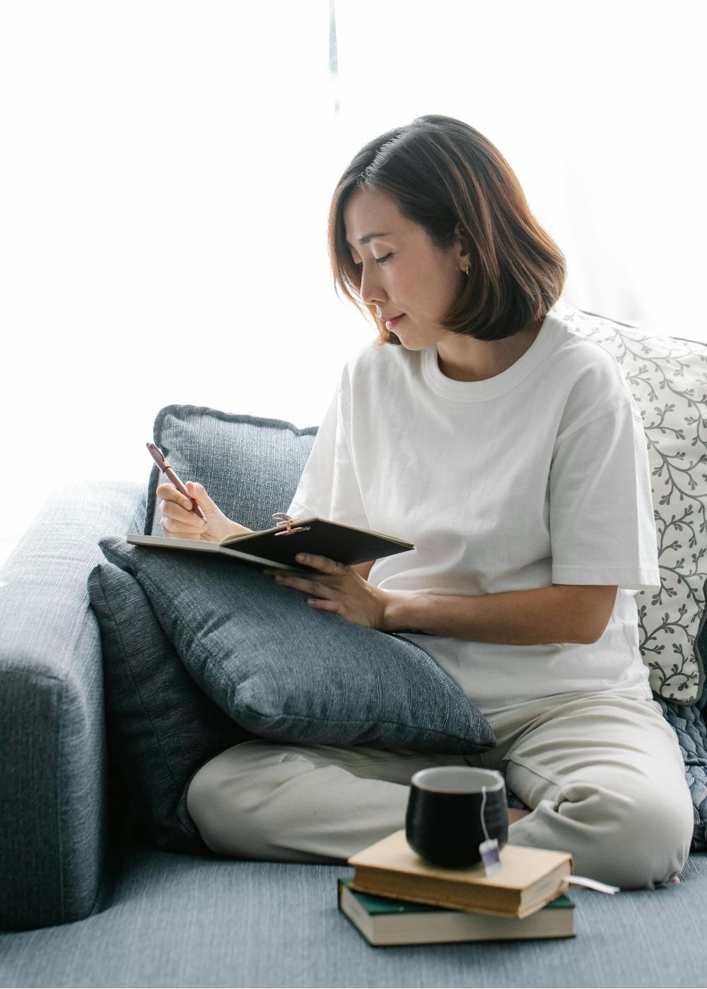 Woman sitting on a gray couch, writing in a notebook with a pen, with books and a mug of tea on a wooden tray in front of her.
