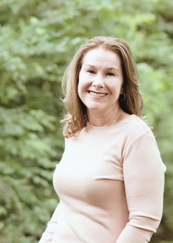 Shannon Champion, Kingwood therapist, wearing a light pink long-sleeved shirt, smiling outdoors, with green foliage in the background.