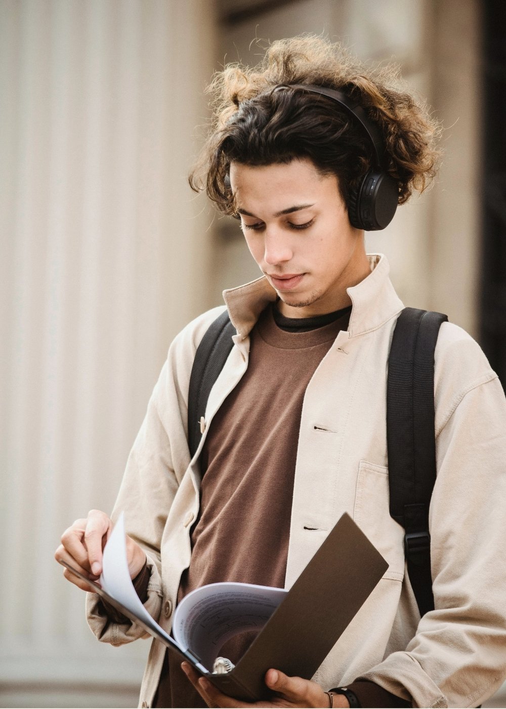 Young man with curly hair wearing headphones, a beige jacket, and a backpack, reading a book outdoors.