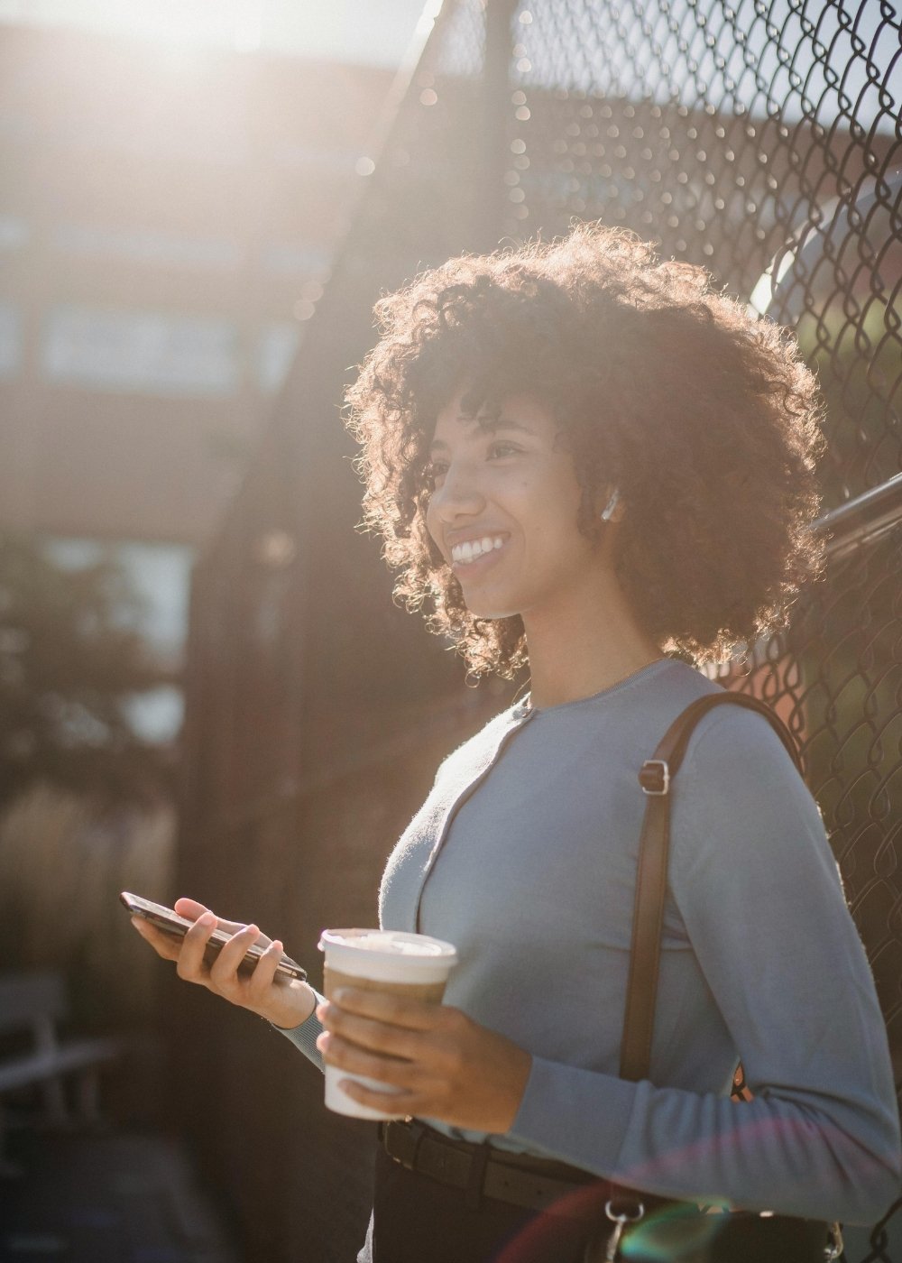 A woman with curly hair smiling and looking to the side while holding a smartphone in one hand and a coffee cup in the other, standing outdoors near a chain-link fence with sunlight shining on her.