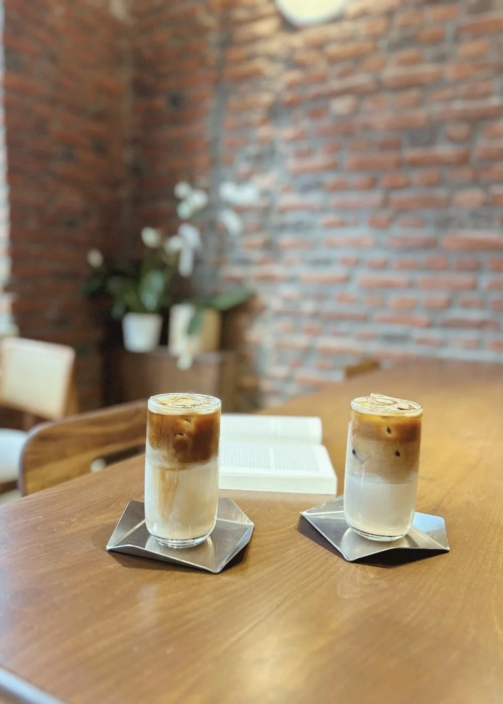 Two glasses of iced coffee with cream and ice cubes on a wooden table, with a book in the background and brick wall decor.