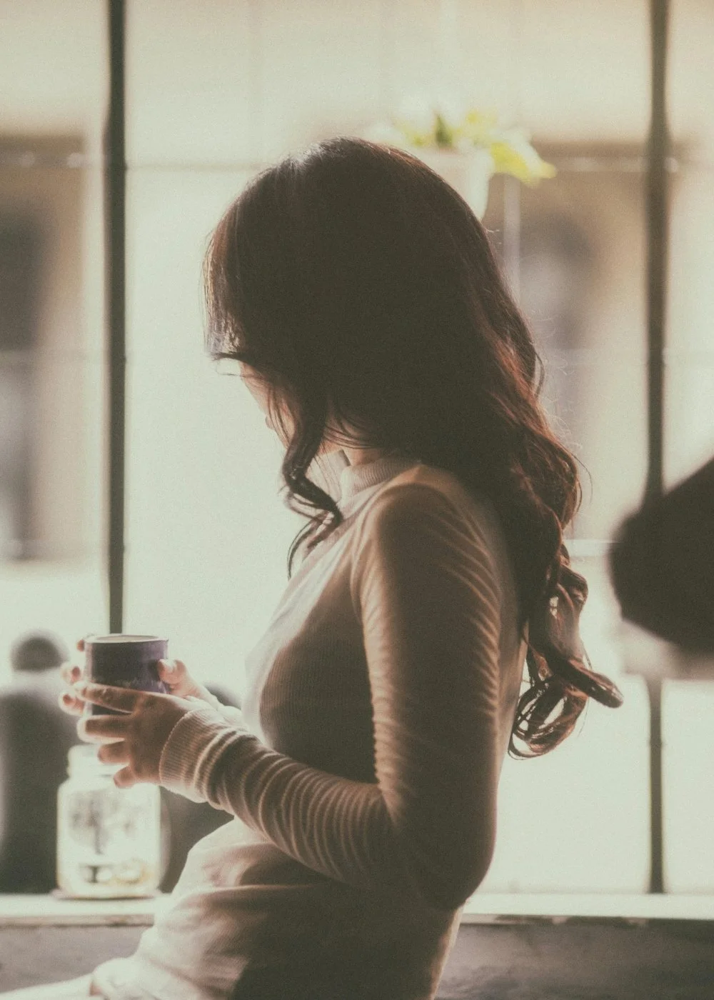 A woman with long dark hair, wearing a beige long-sleeve top, holding a mug, standing indoors looking out window.