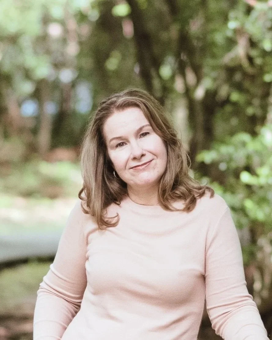 Shannon Champion, Kingwood therapist, wearing a light pink long-sleeved top, smiling outdoors with blurred green foliage in the background.