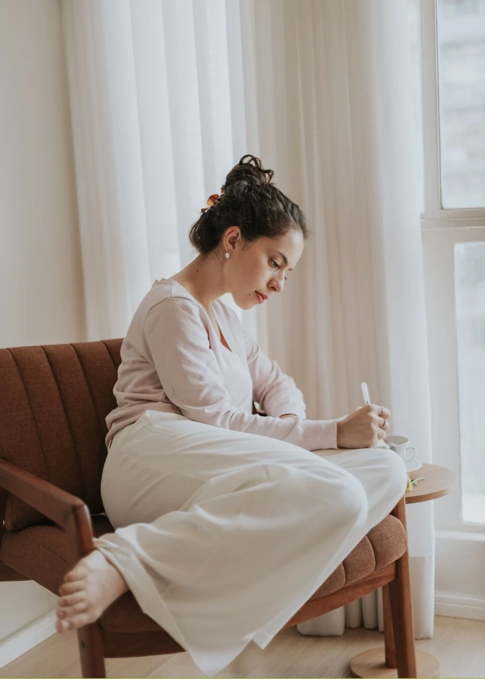 A woman with dark hair styled in a bun and wearing a light pink sweater and white pants, sitting on a brown chair next to a window, writing with a pen in a notebook.