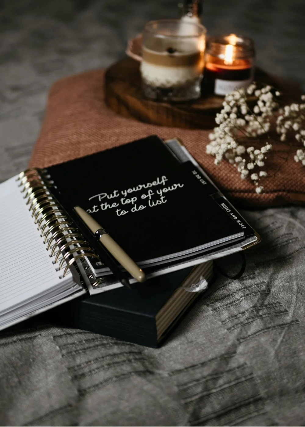 A flat lay of a notebook with the quote 'Put yourself at the top of your to do list' on a black cover, a pen, and a small notepad, all placed on a textured surface with candles and a small bouquet of white baby's breath flowers in the background.