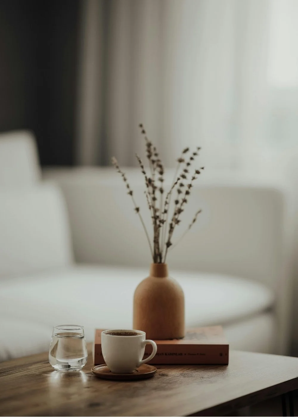 A wooden table with a glass of water, a cup of coffee on a saucer, and a brown vase with dried branches. In the background, a white sofa and light-colored curtains are visible.