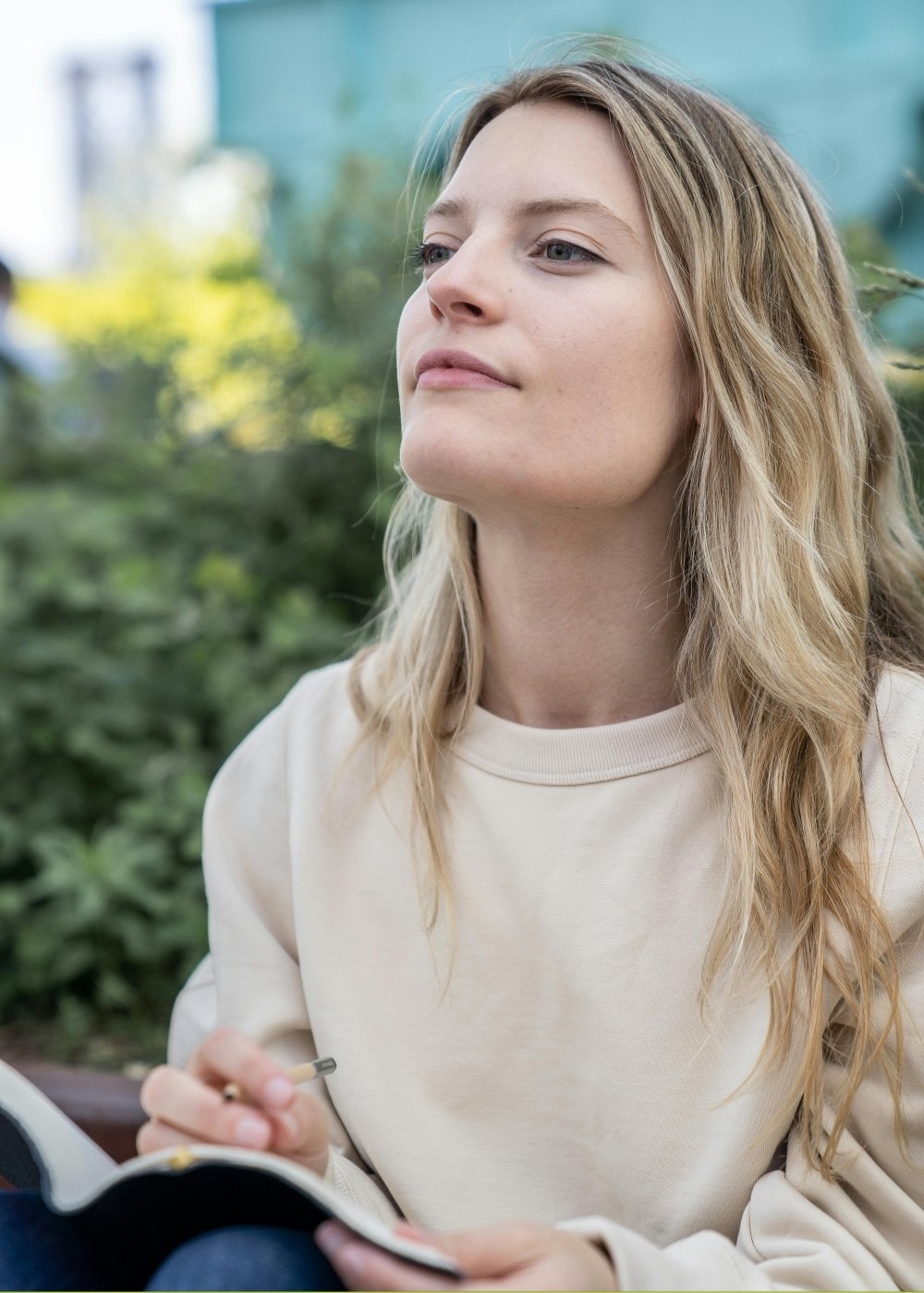 A young woman with blond hair sitting outdoors, holding a pen and open notebook, looking thoughtful.
