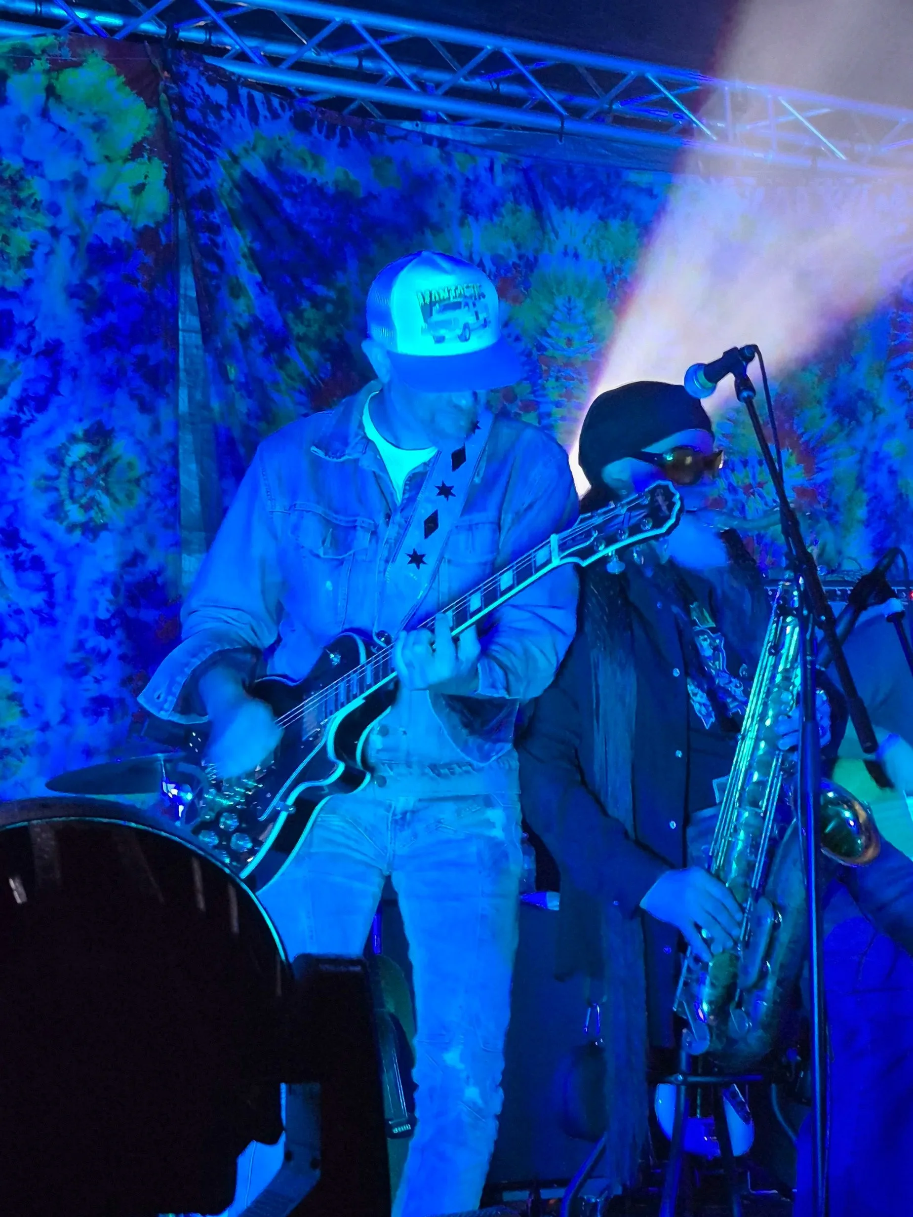 Musicians performing on stage with colorful, tie-dye background, one playing guitar and another on saxophone, illuminated with blue stage lighting.
