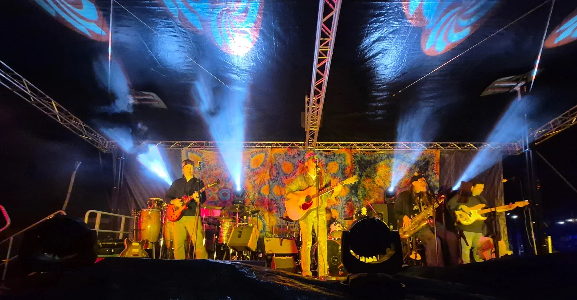 A band performing on a stage at night, with colorful lights and a tie-dye backdrop, featuring musicians playing guitars, drums, and other instruments.