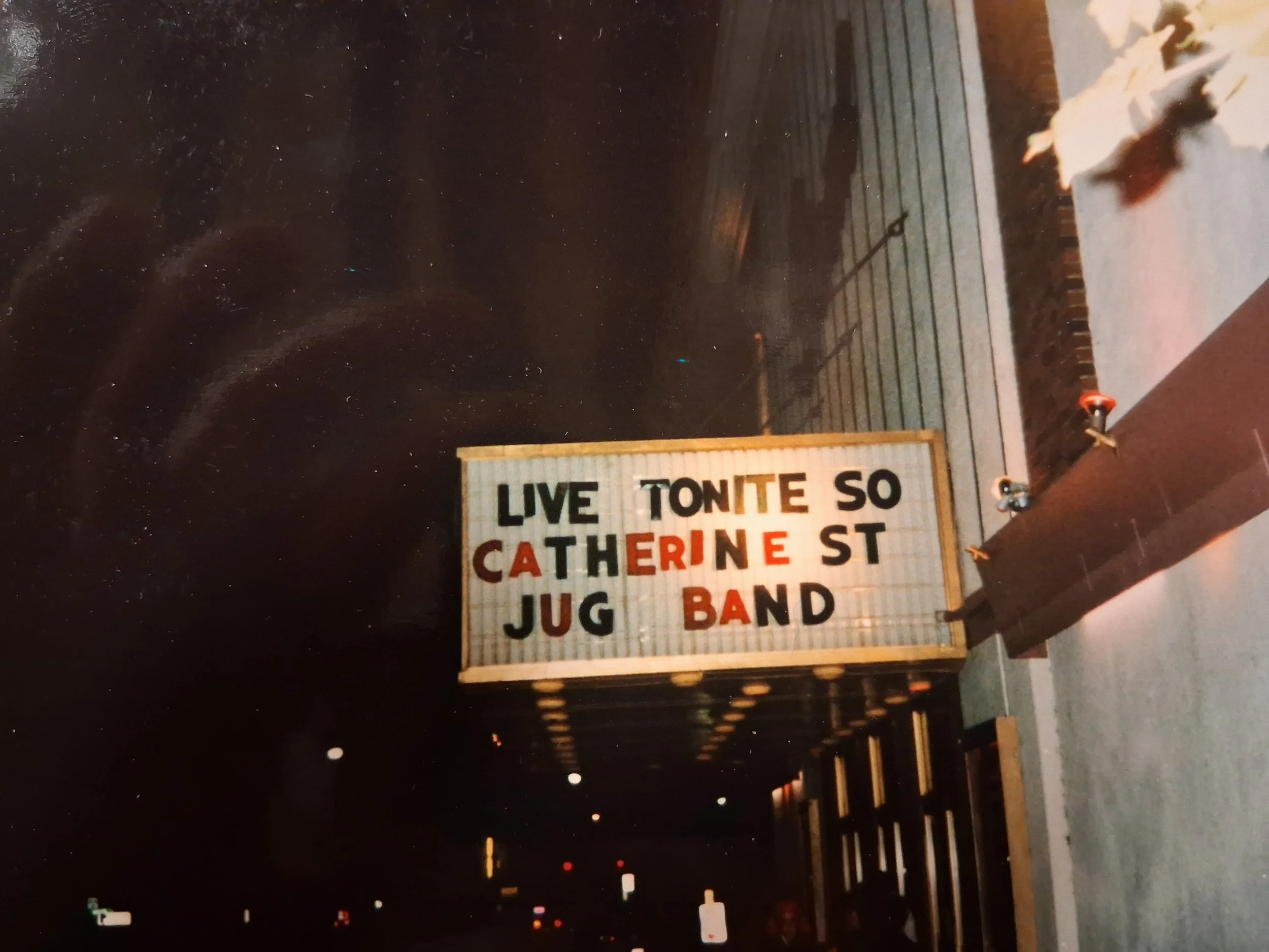 A sign on a building listing 'Live Tonight South Catherine St Jug Band.' The building is at night with a dark sky and small lights in the background.