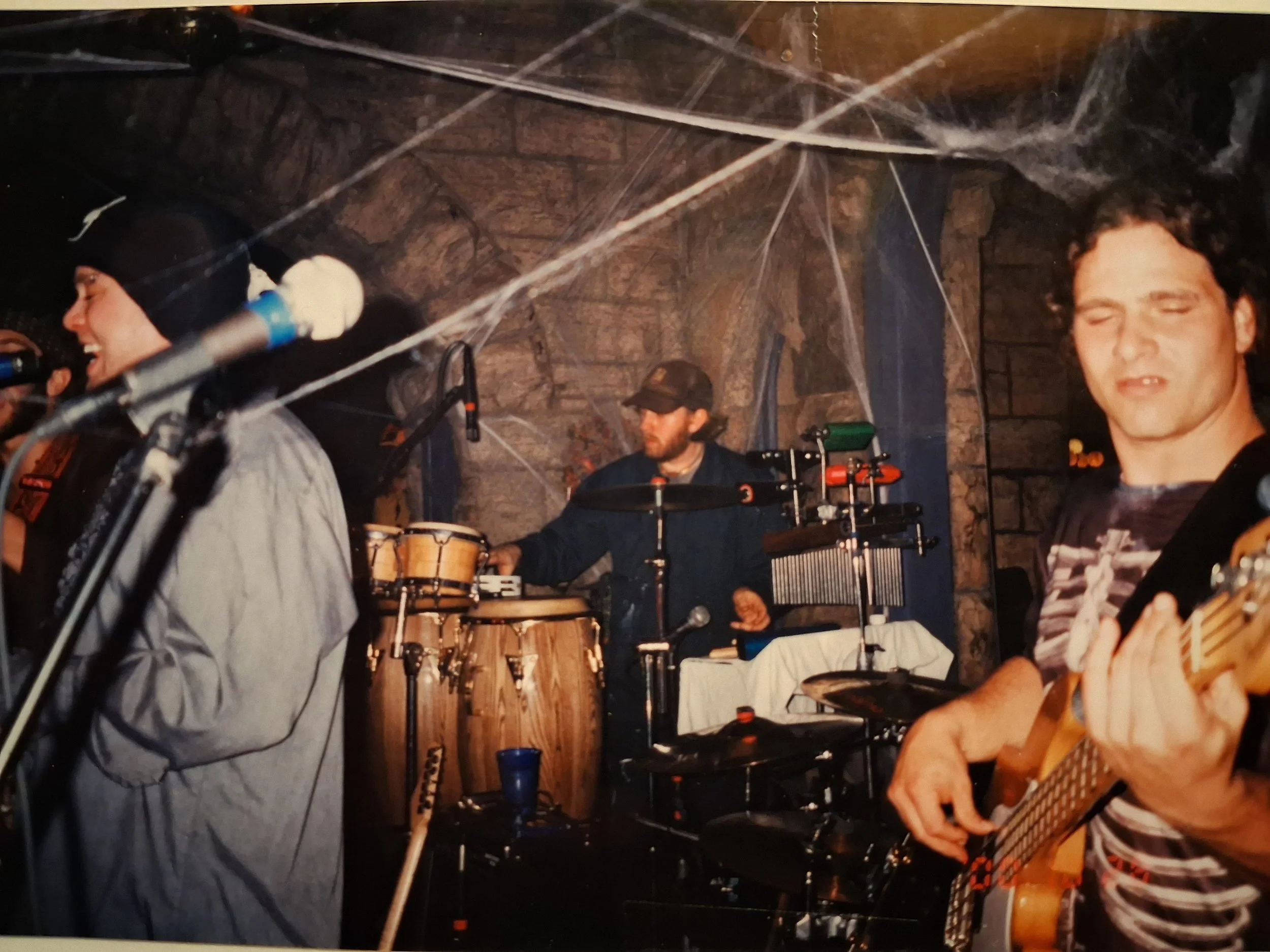 A band performing on stage, with musicians playing various instruments including a guitar and percussion, in a venue with brick walls and cobwebs.