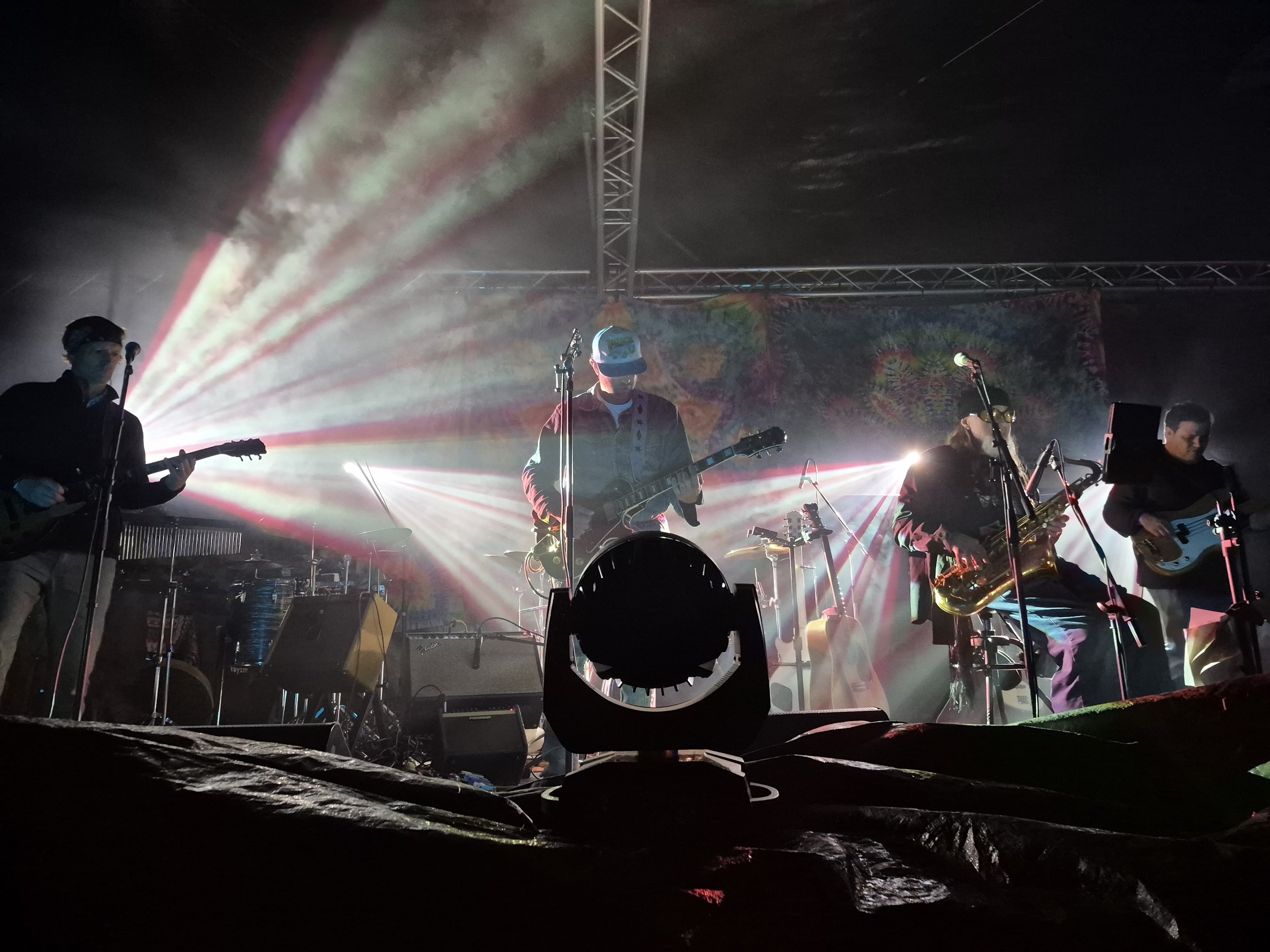 A band performing on stage at night with vibrant lights, including a guitarist and saxophonist, facing away from the audience with a colorful tie-dye backdrop.