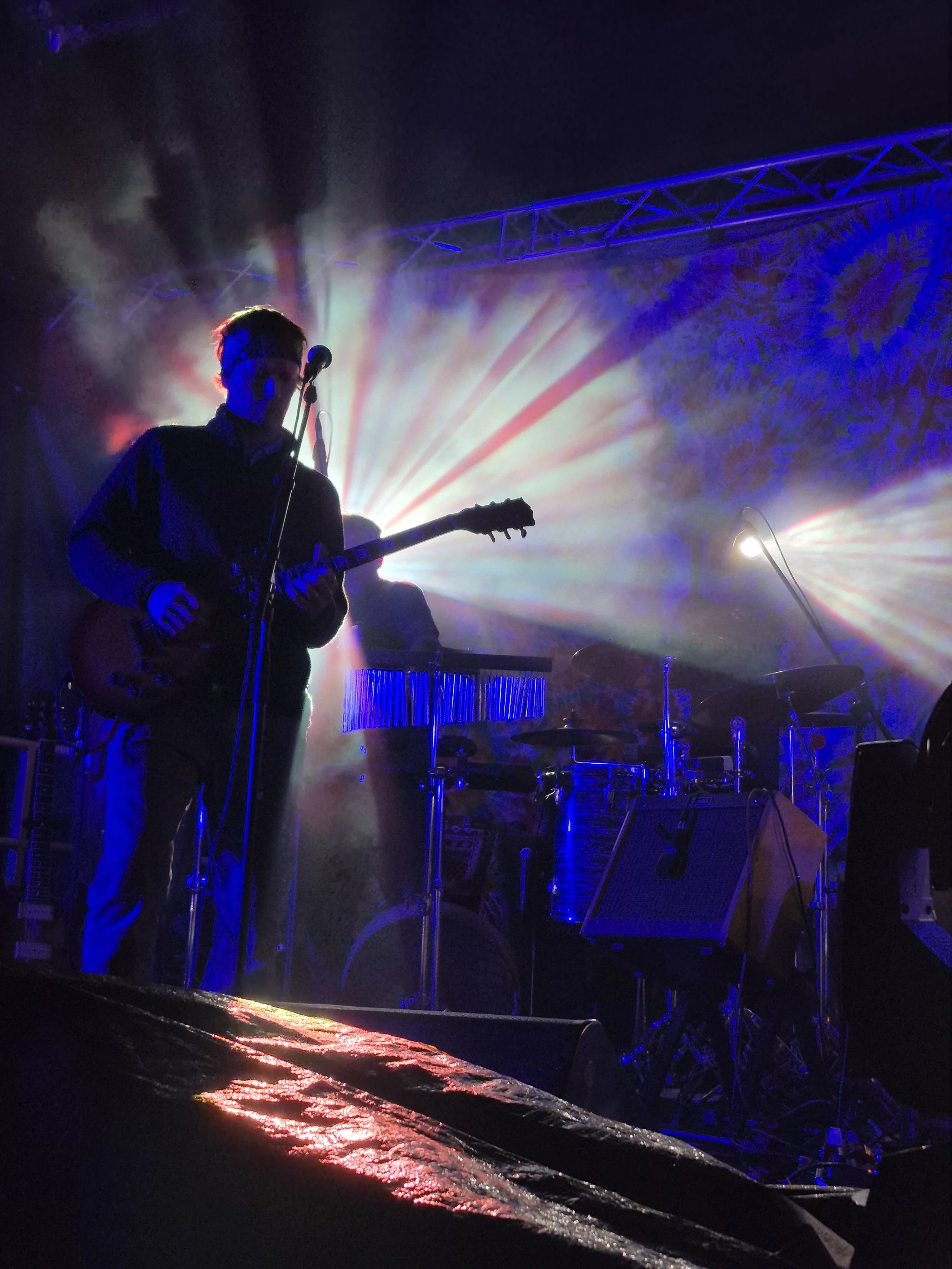 A musician playing guitar on stage with colorful lighting and fog effects, with a silhouette of his face visible, in a concert setting.