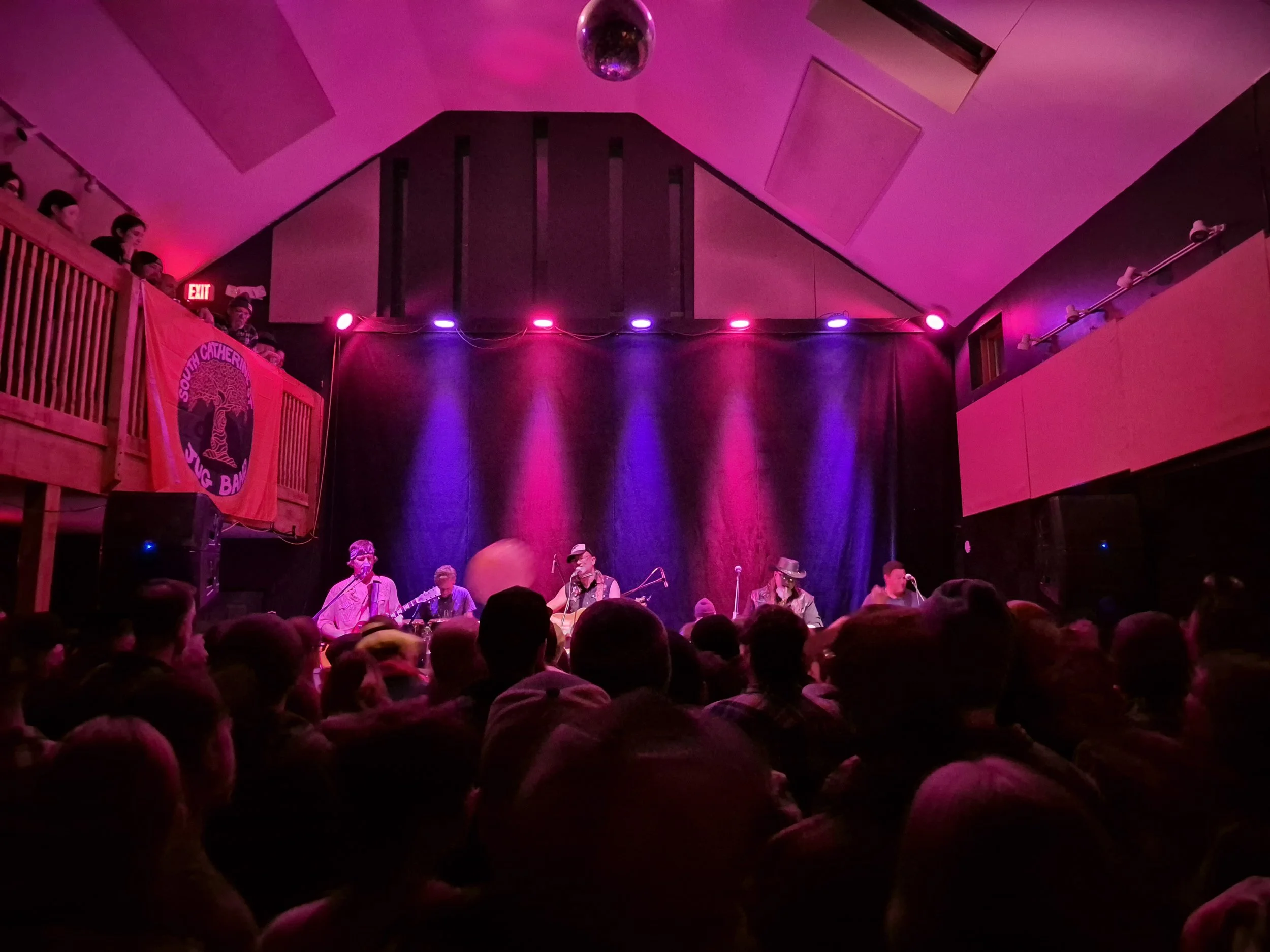 A live music performance on stage with a band playing, illuminated by pink and purple stage lights, with an audience in the foreground.