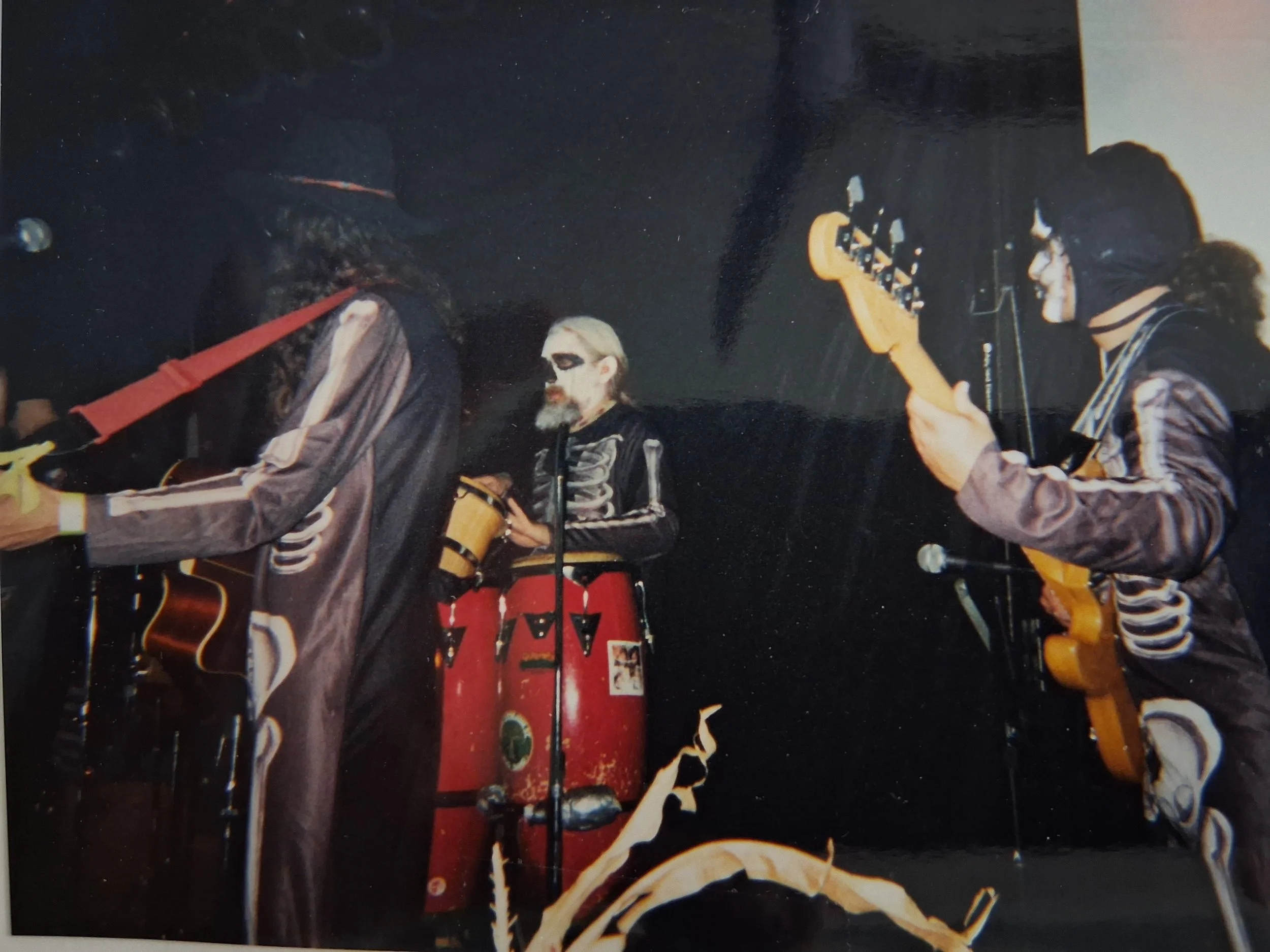 Musicians performing on a dark stage with guitars and a red drum set.