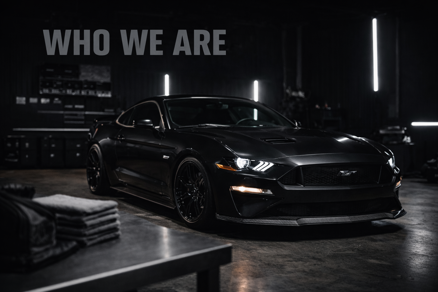 Black sports car in a garage with the words 'WHO WE ARE' written on the wall.